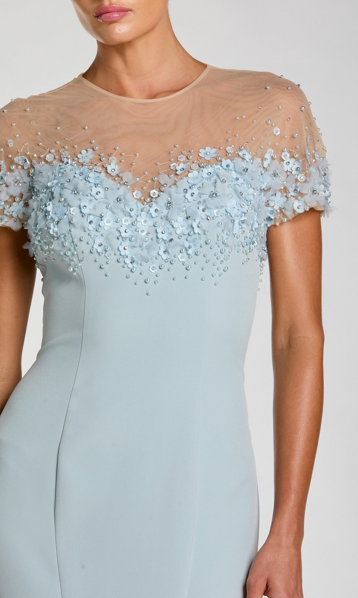 A woman wears the Mac Duggal 11867 short sleeve long formal dress in light blue, featuring a sheer nude mesh top embellished with small blue flowers and beads; only her upper body is visible.